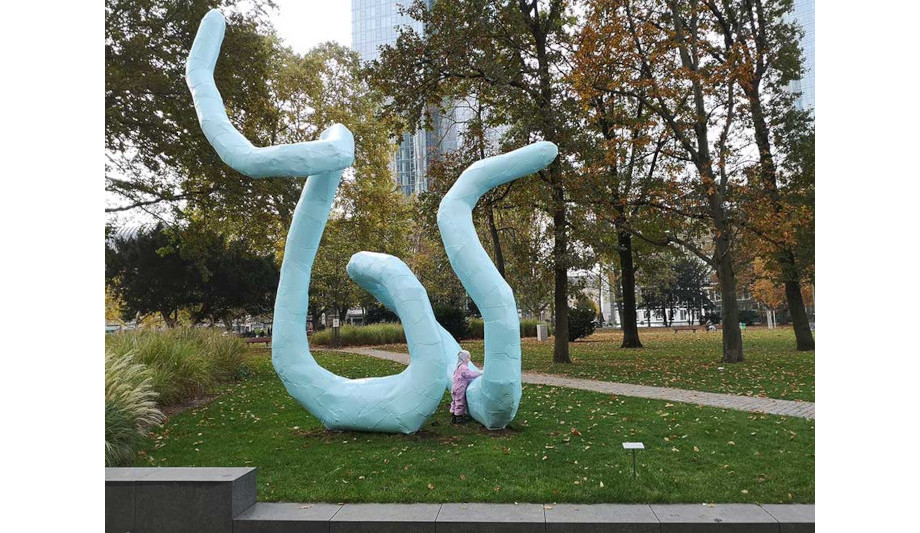 In einem Park mit Wiese und großen Bäumen steht eine große blaue Skulptur aus Aluminium. Ihre Form gleicht der einer riesigen Schlange. Ein Kind stützt sich auf die Skulptur.