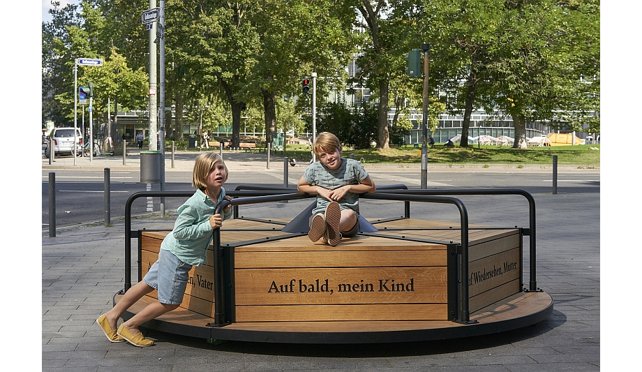 Auf einem breiten Fußgängerweg an einer Straßenkreuzung steht ein Denkmal in Form eines Karussells. Ein Kind sitzt auf dem Karussell, ein weiteres Kind schiebt es mit aller Kraft an. Drei Sätze sind in die Seiten aus Holz graviert: „Auf bald, mein Kind“, „Auf Wiedersehen, Mutter“ und „Auf Wiedersehen, Vater“.