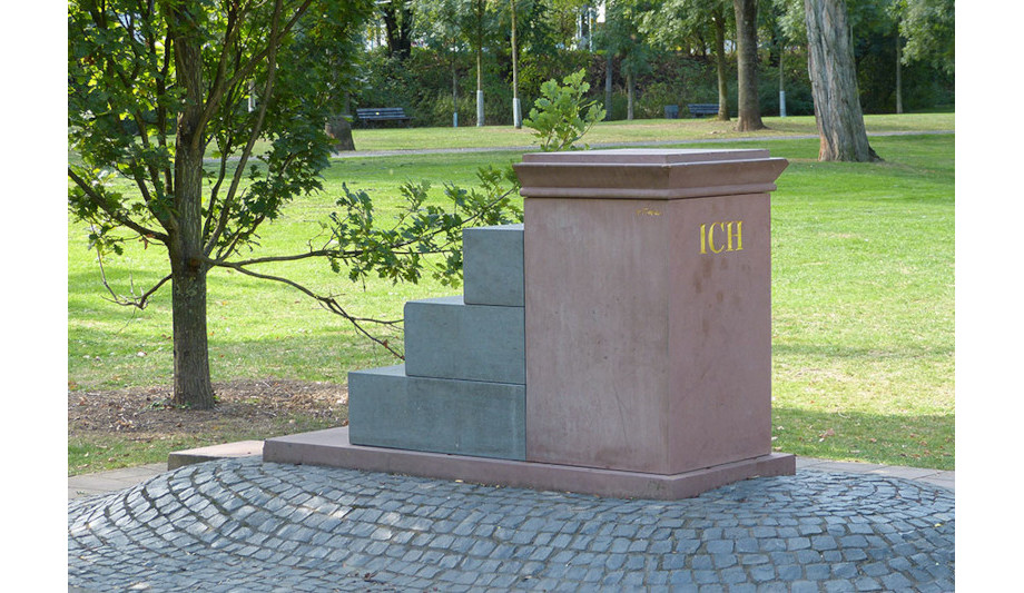 Auf einer Wiese steht ein Sockel aus rötlichem Stein. Das Wort „ICH“ ist in goldenen Buchstaben auf eine Seite geschrieben. An der rückwärtigen Seite befinden sich Stufen aus grauem Stein, die auf den Sockel hinaufführen.