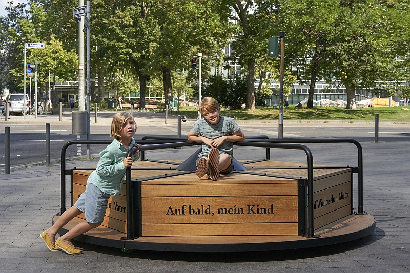 Auf einem breiten Fußgängerweg an einer Straßenkreuzung steht ein Denkmal in Form eines Karussells. Ein Kind sitzt auf dem Karussell, ein weiteres Kind schiebt es mit aller Kraft an. Drei Sätze sind in die Seiten aus Holz graviert: „Auf bald, mein Kind“, „Auf Wiedersehen, Mutter“ und „Auf Wiedersehen, Vater“.