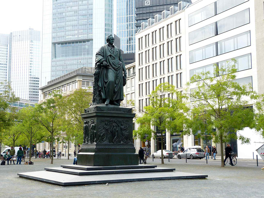 Zu sehen ist das bronzene Denkmal für Johann Wolfgang von Goethe auf dem Goetheplatz. Über einem hohen Sockel erhebt sich die monumentale Statue des Dichters. Er hält eine Schriftrolle und einen Lorbeerkranz. Den Sockel schmücken Reliefs mit Figuren aus Goethes Werken.