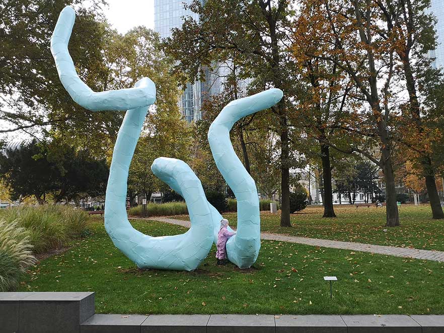 In einem Park mit Wiesen und Bäumen steht eine große blaue Skulptur aus Aluminium. Ihre Form gleicht der einer riesigen Schlange. Ein Kind stützt sich auf die Skulptur.