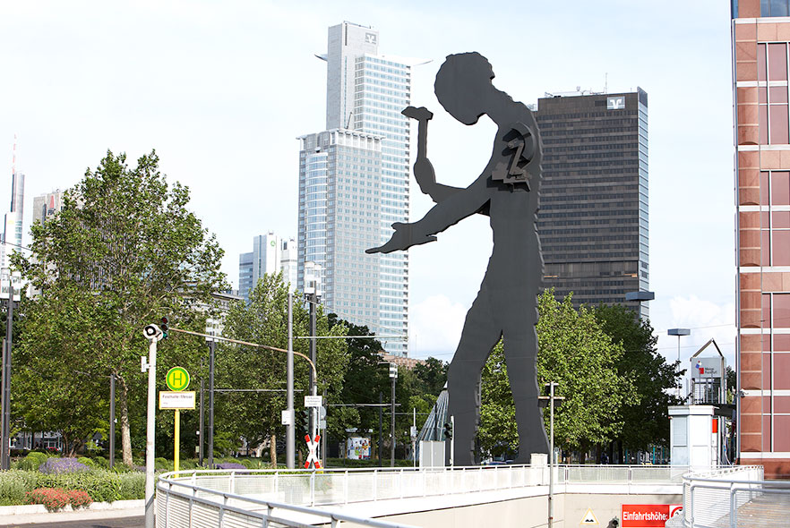 An einer breiten Straße am Messegelände steht die gigantische, schwarze Silhouette eines Menschen. In der einen Hand hält sie einen Gegenstand, in der anderen einen Hammer. Auf Schulterhöhe ist die Maschinerie angebracht, die den Arm mit dem Hammer auf und ab bewegt.