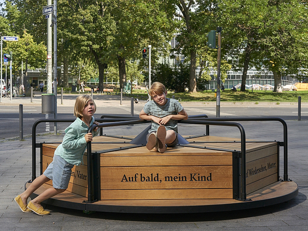 Auf einem breiten Fußgängerweg an einer Straßenkreuzung steht ein Denkmal in Form eines Karussells. Ein Kind sitzt auf dem Karussell, ein weiteres Kind schiebt es mit aller Kraft an. Drei Sätze sind in die Seiten aus Holz graviert: „Auf bald, mein Kind“, „Auf Wiedersehen, Mutter“ und „Auf Wiedersehen, Vater“.