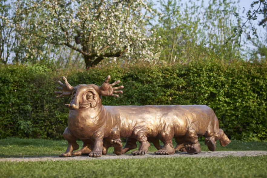 Mitten im Grün steht ein wurmartiges Fabelwesen aus Bronze. Es läuft auf sechs Paar Füßen. Auch seine Nase und sein Schwanz sind wie Füße geformt, seine Ohren allerdings wie Hände. Mit seinem Lächeln strahlt das Wesen Freundlichkeit aus.