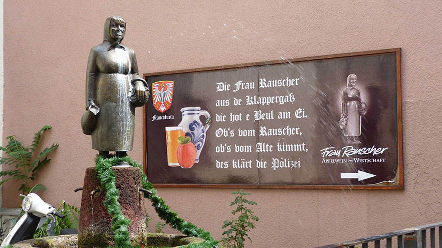 Auf einem Brunnen steht die Bronzefigur der Fraa Rauscher. Sie trägt ein Kopftuch und führt einen Krug und einen Korb mit. Im Hintergrund wirbt ein Schild für ein Apfelwein-Lokal, unter anderem ist hier ein Ausschnitt aus dem Apfelweinlied über die Fraa Rauscher zu lesen.